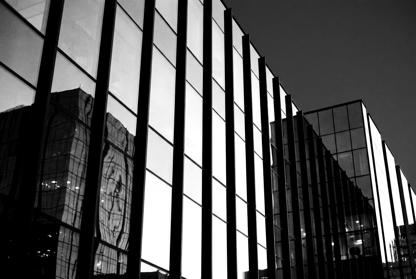A modern glass facade at dusk reflecting another building, in black and white